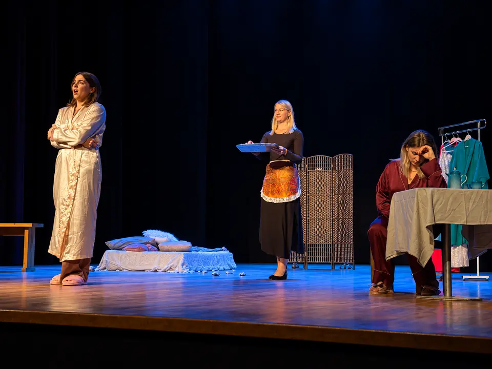 Three performers in a scene with a bed and table